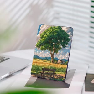 Könnte beinhalten: Ein rechteckiger Handyhalter mit einer Landschaftsillustration. Das Kunstwerk zeigt einen großen grünen Baum in einem Feld aus goldenem Weizen, mit einer Bank, einem kleinen Haus und Bergen unter einem bewölkten blauen Himmel. Der Halter liegt auf einer weißen Oberfläche.