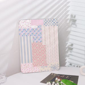 May include: A rectangular decorative panel with a patchwork design. The panel features various floral and striped patterns in shades of pink, blue, and white. The panel is leaning against a white wall.