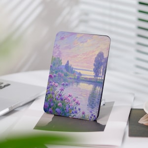 Può includere: Un dipinto che rappresenta una scena di lago con fiori viola in primo piano, alberi sulla riva e un cielo di tramonto rosa e arancione.