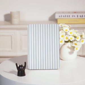 May include: A rectangular, light blue and white striped notebook stands upright on a white table. A small, black cat figurine with raised arms is in front of the notebook. A white vase with white and yellow flowers is to the right.