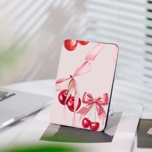 Könnte beinhalten: Eine rechteckige Handyhülle mit einem Aquarelldesign aus Kirschen und rosa Schleifen auf hellrosa Hintergrund. Die Kirschen sind tiefrot und die Schleifen sind zartrosa. Die Hülle hat einen schwarzen Rand.