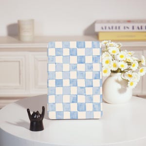 Könnte beinhalten: Ein rechteckiges Notizbuch mit blau-weißem Karomuster steht auf einem weißen Tisch. Vor dem Notizbuch steht eine kleine schwarze Katzenfigur mit erhobenen Armen. Rechts befindet sich eine weiße Vase mit weißen und gelben Blumen.