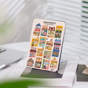 Peut inclure: Une feuille d'autocollants colorés illustrant différentes devantures de magasins. Les autocollants sont disposés en grille et comprennent des boutiques comme "Cozy Bakery", "Flower Shop" et "Garden Center". La feuille est intitulée "Shopfronts by AnneliesDraws".