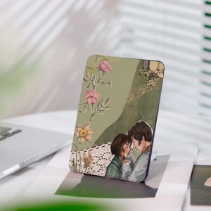 Può includere: Un supporto per telefono rettangolare con un'illustrazione romantica. Il design include una coppia che si abbraccia, elementi floreali con fiori rosa e gialli e un bordo in pizzo. Lo sfondo ha una combinazione di colori verde e oro.