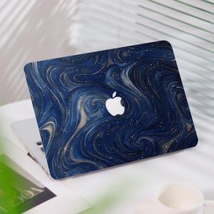 Op de afbeelding: Een laptop met een marineblauwe hoes met een wervelend, abstract ontwerp met gouden en witte accenten. Het Apple-logo is zichtbaar. De laptop staat open op een wit oppervlak.