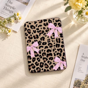 May include: A rectangular e-reader case featuring a tan and black leopard print pattern decorated with three light pink bows. The case has rounded corners and a smooth surface, displaying a repeating animal skin design across the entire front cover.