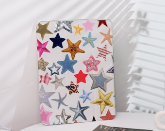 Può includere: Una custodia bianca per tablet ornata da una varietà di disegni a stella colorati. Le stelle sono in vari colori, trame e dimensioni, tra cui denim, paillettes e tessuti fantasia. La custodia è su una superficie bianca.