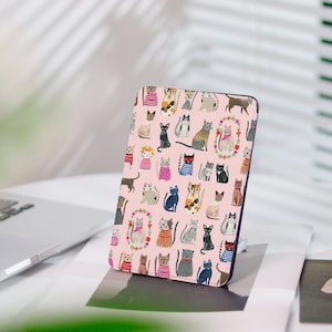 May include: A pink tablet case featuring a repeating pattern of illustrated cats in various poses and outfits. The cats are in different colors and patterns, with some wearing sweaters and floral wreaths. The case is displayed on a white surface.
