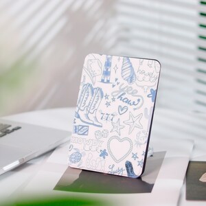 Peut inclure: Un étui de téléphone rectangulaire avec un fond blanc et un motif de griffonnage bleu. Le motif comprend des bottes de cowboy, des cœurs, des étoiles, un phare et le mot "Howdy". L'étui est posé verticalement sur une surface blanche.