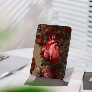 Può includere: Custodia per telefono rettangolare con un'illustrazione dettagliata di un cuore umano nei toni del rosso, su uno sfondo di rose e fogliame. Il design ha uno stile vintage e artistico, con un focus sui dettagli anatomici e sugli elementi floreali.