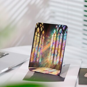 May include: A rectangular phone stand featuring a vibrant stained-glass window design. The image showcases a colorful array of light streaming through the window, creating a rainbow effect. The stand is propped up on a white surface.