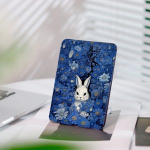 Könnte beinhalten: Rechteckige Handyhülle mit einem Design eines weißen Kaninchens inmitten blauer Blumen. Die Illustration hat einen skurrilen Stil. Die Hülle steht auf einer weißen Oberfläche.