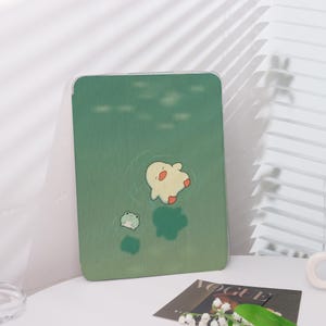 Op de afbeelding: Groene tablethoes met een cartoon eend die in het water drijft. De eend is geel met rode voeten. Er is ook een kleine groene kikker afgebeeld. De hoes heeft een doorzichtige rand en ligt op een wit oppervlak.