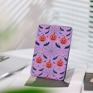 May include: A rectangular phone case with a Halloween design. It features orange pumpkins, black bats, spiderwebs, and candles on a lilac background. The case is on a white surface.