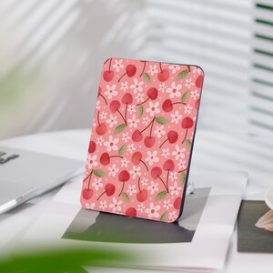 Peut inclure: Une housse rose pour liseuse avec un motif de cerises et de fleurs. La housse présente un motif répétitif de cerises rouges avec des tiges vertes et des fleurs blanches.