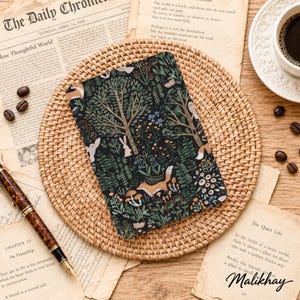 Op de afbeelding: Een rechthoekig notitieboek met een donker bospatroon met illustraties van vossen, konijnen, muizen, bomen, paddenstoelen en diverse planten. De kaft heeft een matte afwerking en ligt centraal op een ronde placemat van gevlochten rotan.
