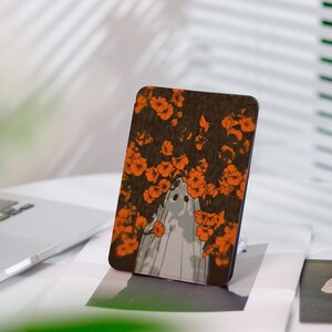 Può includere: Un supporto per telefono rettangolare con un design a fantasma. Il fantasma è circondato da fiori arancioni su uno sfondo marrone scuro. Il supporto è appoggiato su una superficie bianca, ideale per tenere un telefono.