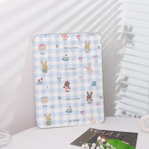 May include: A light blue and white checkered tablet case with cartoon bears, bunnies, and cakes. The case has the text "Hello my friend" and musical notes. The case is on a white surface.