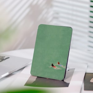 May include: A green rectangular case featuring an illustration of two people swimming in water. The case has a black border and is resting on a white surface. A laptop is visible in the background.
