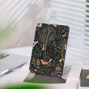 Könnte beinhalten: Eine rechteckige Handyhülle mit einer skurrilen Waldszene. Das Design zeigt Bäume, Füchse, Kaninchen, Eulen und Pilze in Grün-, Braun- und Orangetönen auf schwarzem Hintergrund. Die Hülle ist abgewinkelt, um die detaillierte Grafik hervorzuheben.