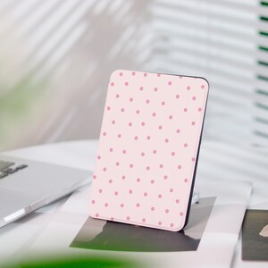 Puede incluir: Un lector electrónico rectangular con una cubierta de color rosa claro adornada con un patrón repetitivo de pequeños puntos de color rosa más oscuro. El dispositivo tiene un marco negro delgado y está colocado verticalmente.