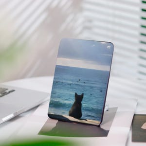 Könnte beinhalten: Rechteckige Handyhülle mit einer ruhigen Ozeanszene und einer schwarzen Katzen-Silhouette. Die Katze blickt unter einem bewölkten Himmel auf das Wasser. Die Hülle liegt auf einer weißen Oberfläche und deutet auf ein Küsten- oder Naturthema hin.