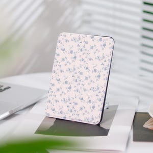 Könnte beinhalten: Eine weiße E-Reader-Hülle mit einem blauen Blumenmuster. Die Hülle ist aus einem weichen, strapazierfähigen Material gefertigt und wurde entwickelt, um Ihren E-Reader vor Kratzern und Stößen zu schützen.