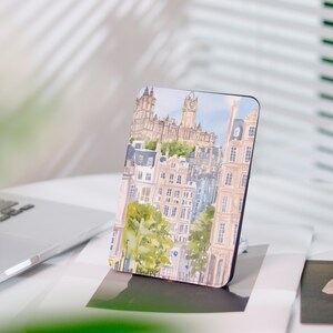 May include: A rectangular phone stand with a watercolour painting of a European cityscape, including buildings, trees, and a clock tower. The colour scheme features beige, blue, and green hues. The stand is placed on a white surface.