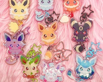 Pode incluir: Uma coleção de chaveiros coloridos de personagens de desenhos animados. Os chaveiros apresentam várias criaturas com olhos grandes e designs exclusivos, presos a chaveiros de metal e pingentes em forma de estrela. O fundo é uma superfície macia e rosa.