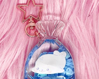 May include: A clear, bag-shaped keychain with a blue liquid interior and a white seal illustration. The keychain has a gold star-shaped clasp. The background is pink, fluffy fabric.