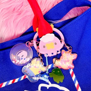 May include: A blue tote bag with a red strap and a collection of colourful keychains. The keychains include a pink cartoon character, a clear circular charm with a character inside, and other animal-shaped charms.
