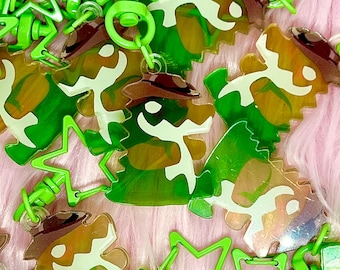 May include: A collection of green and brown dinosaur-shaped keychains with a cowboy hat design. The keychains are made of translucent plastic and have a star-shaped clip. The background is a soft pink.