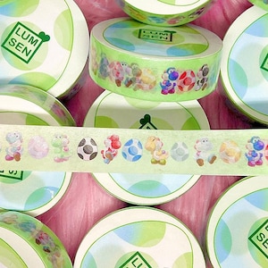 Könnte beinhalten: Grünes und weißes Washi-Tape mit einem sich wiederholenden Muster aus Comicfiguren und Eiern. Das Band ist aufgerollt und auf einer rosa Oberfläche mit anderen Bandrollen ausgestellt. Das Band ist mit "Lum Sen" beschriftet.