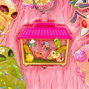 May include: A pink plastic charm with a clear plastic window showing a cartoon image of a house with a roof and a yellow, green, and blue insect inside. The charm is attached to a silver key ring.