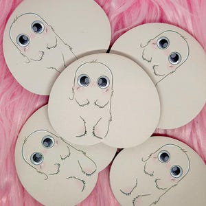 May include: Five circular stickers featuring a light beige, furry creature with large, dark, expressive eyes and small hands. Each sticker displays the same illustration of the creature with a soft, rounded body, subtle pink blush on its cheeks, and a simple, clean outline, presented in a group arrangement.