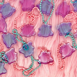 May include: A collection of colorful, translucent charm necklaces. Each charm is a stylized, amorphous shape in shades of pink and purple, with a smiling face. The necklaces feature pink and teal beaded chains, creating a playful and vibrant aesthetic.