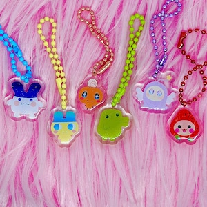 May include: A collection of colorful acrylic charms featuring various cartoon characters. Each charm is attached to a beaded chain in a coordinating color. The charms are in the shape of a bunny, a creature with a swirl on its head, a green creature, a winged creature, and a strawberry.