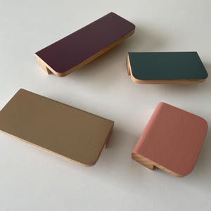Puede incluir: Cuatro tiradores rectangulares para armarios en tonos ciruela, verde, beige y coral. Cada tirador tiene una parte superior pintada y una base de madera natural. Los tiradores están dispuestos sobre una superficie blanca, mostrando su diseño moderno y variaciones de color.