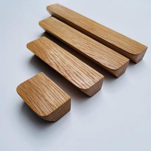 Op de afbeelding: Vier houten kastgrepen met een natuurlijke houtnerf afwerking. De grepen zijn rechthoekig met afgeronde randen en zijn diagonaal op een wit oppervlak geplaatst. Het hout heeft een warme, lichtbruine kleur.