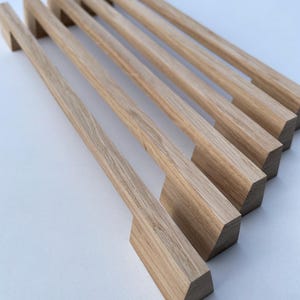 Pode incluir: Seis puxadores de armário de madeira com acabamento natural de grão de madeira. Cada puxador tem uma pega retangular e uma base quadrada. Os puxadores estão dispostos diagonalmente, mostrando o seu design e textura. A madeira clara proporciona uma estética limpa e moderna.
