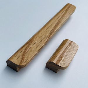 Può includere: Due maniglie per mobili in legno. Una è di forma rettangolare e allungata, l'altra è più piccola e curva. Entrambe sono realizzate in legno marrone chiaro con venature visibili.