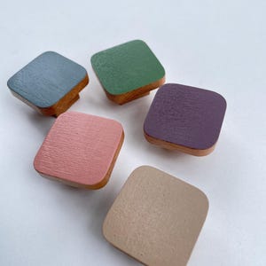 Op de afbeelding: Vijf vierkante houten kastknoppen in verschillende kleuren: blauw, groen, paars, roze en beige. Elke knop heeft een geverfde bovenkant en een natuurlijke houten basis. De knoppen zijn gerangschikt op een witte ondergrond.