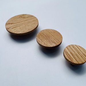 May include: Three wooden cabinet knobs in varying sizes. The knobs are round with a flat top and a short, cylindrical base. The wood grain is visible, and the knobs are a light brown color. They are arranged diagonally on a white surface.