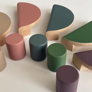 Może przedstawiać: Kolekcja drewnianych gałek i uchwytów do szafek w różnych kształtach i kolorach. Gałki są cylindryczne, a uchwyty półokrągłe. Kolory obejmują beżowy, koralowy, morski, zielony i fioletowy.