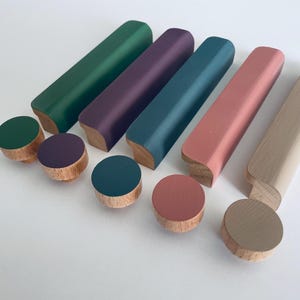 Puede incluir: Una colección de seis tiradores y pomos de madera para armarios en varios colores. Los tiradores son rectangulares con bordes redondeados, pintados en verde, morado, turquesa, rosa y madera natural. Los pomos son redondos con base de madera y tapas pintadas a juego.