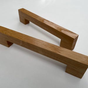 Könnte beinhalten: Zwei rechteckige Holzschrankgriffe mit natürlicher Eichenholzoberfläche. Die Griffe haben ein schlichtes, modernes Design mit einem rechteckigen Griff und zwei quadratischen Montagepfosten. Ideal für Küchen- oder Badezimmerschränke.