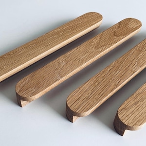 Puede incluir: Un juego de cuatro tiradores de madera de color marrón claro. Cada tirador es rectangular con extremos redondeados y un pequeño labio curvado en un lado. La veta de la madera es visible.