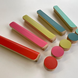 Pode incluir: Uma coleção de puxadores e botões de armário de madeira coloridos. Os puxadores são retangulares com bordas arredondadas, pintados em vermelho, rosa, amarelo, azul-turquesa e azul. Os botões são redondos e pintados para combinar. O grão da madeira é visível.