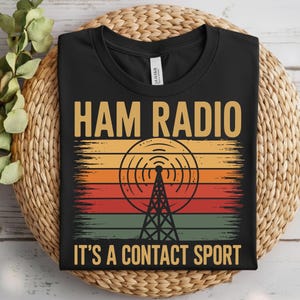 Peut inclure: T-shirt noir avec l'inscription "HAM RADIO" en lettres dorées au-dessus d'un graphique de tour radio et d'ondes. En dessous, le texte indique "IT'S A CONTACT SPORT". Le t-shirt est présenté sur un panier en osier.