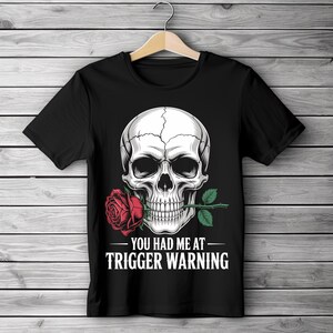 May include: Black t-shirt featuring a detailed skull graphic holding a red rose in its teeth. The text "YOU HAD ME AT TRIGGER WARNING" is printed below the skull in white. The design is centered on the shirt.